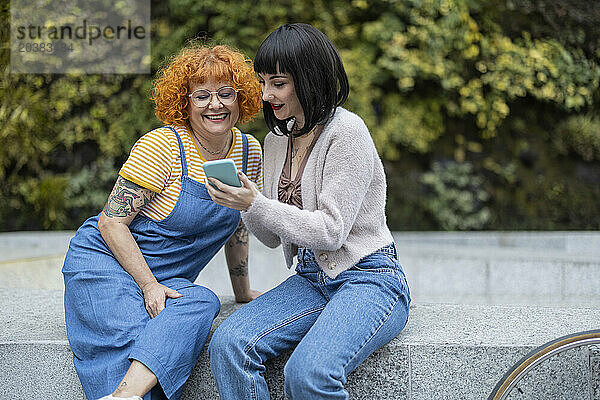 Daughter sharing smart phone with mother sitting on retaining wall