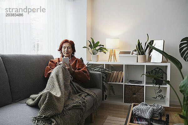 Retired senior woman using smart phone with headphones sitting on couch in living room