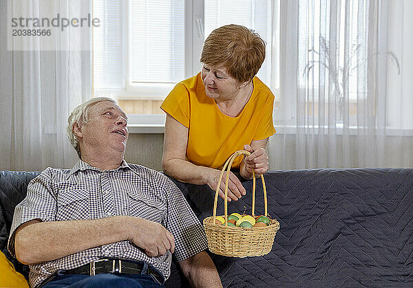 Senior woman holding Easter basket and talking to man sitting on sofa at home