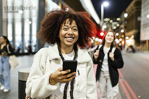 Happy young woman holding smart phone and standing on street