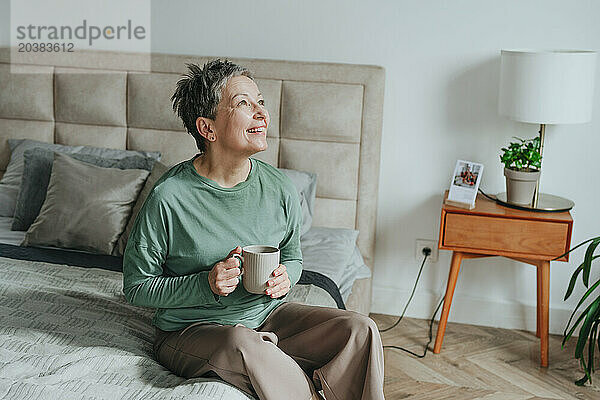 Happy woman holding coffee mug sitting on bed at home