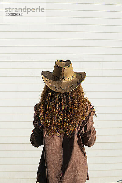 Curly haired woman wearing jacket and cowboy hat in front of white shutter