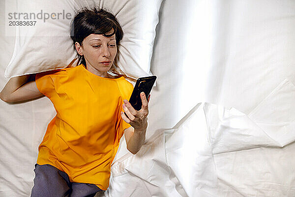 Woman in yellow t-shirt lying with phone on bed at home