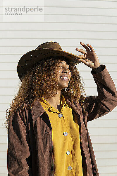 Smiling young woman with eyes closed wearing jacket and cowboy hat on sunny day