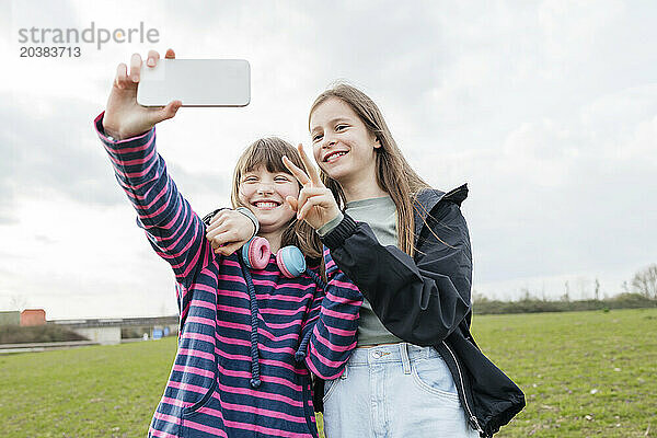 Happy girl taking selfie with friend showing peace sign at lawn