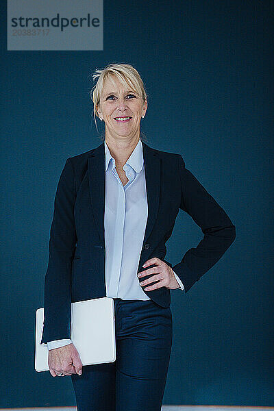 Happy businesswoman standing with laptop against navy blue background