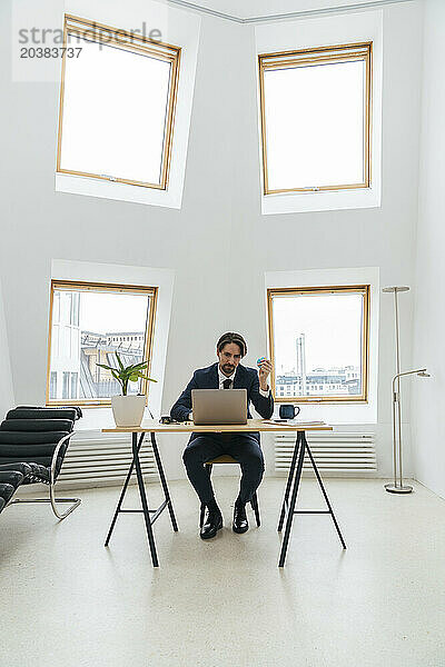 Businessman with globe using laptop sitting at desk in front of windows