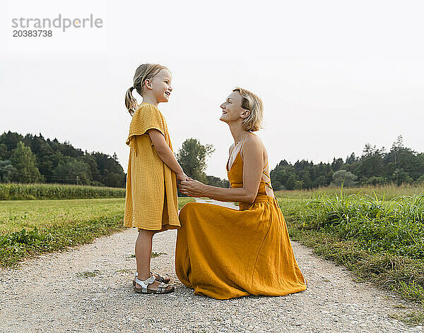 Mother holding hands of daughter and crouching on footpath near meadow