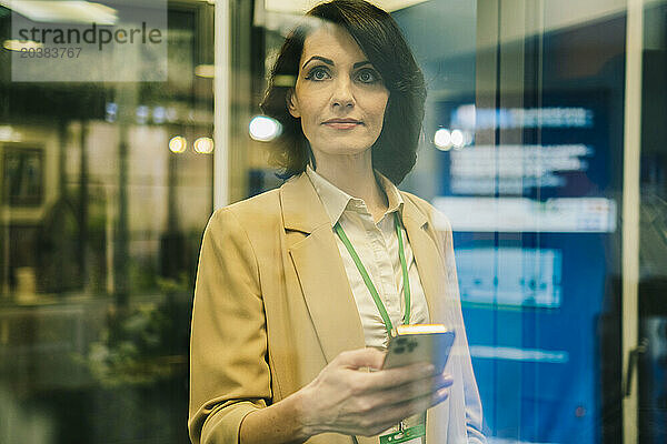 Confident mature businesswoman with smart phone seen through glass at office