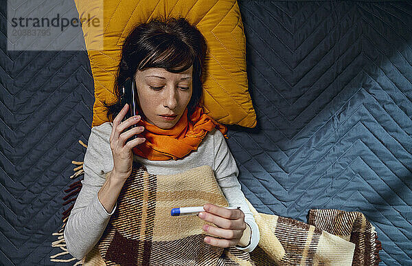 Woman talking on smart phone and holding thermometer lying on bed at home