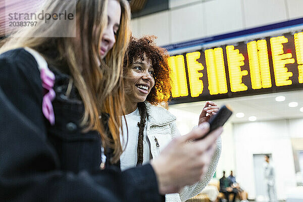 Happy woman with friend using smart phone at railroad station