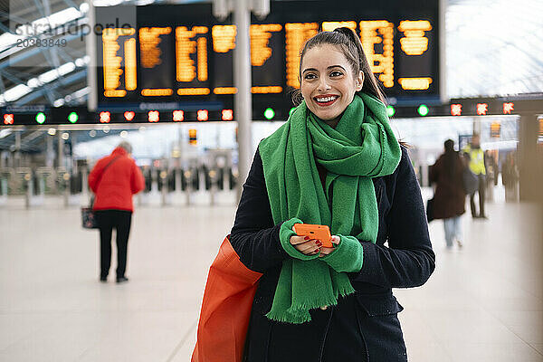 Happy woman wearing green scarf holding smart phone at train station