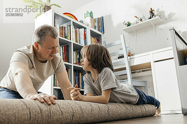 Father talking to boy lying on rolled up carpet at home