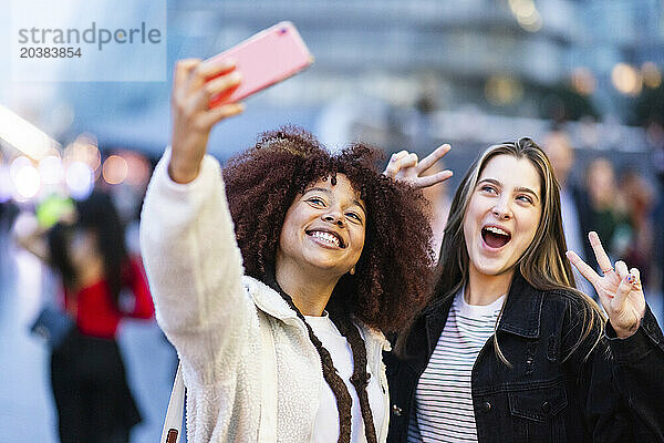 Cheerful woman taking selfie with friend gesturing peace sign