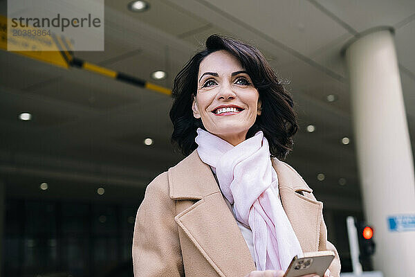 Happy beautiful mature businesswoman wearing overcoat and scarf standing with smart phone