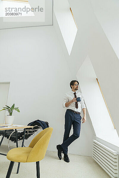 Businessman with coffee cup leaning on wall looking out through window at office