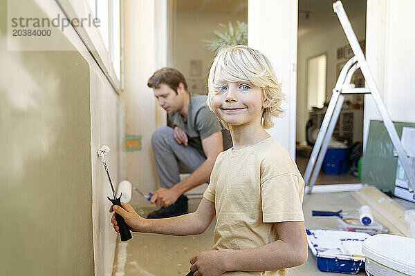 Smiling boy painting wall with roller near father at home