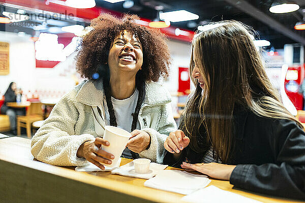 Cheerful woman holding disposable cup with friend in cafe