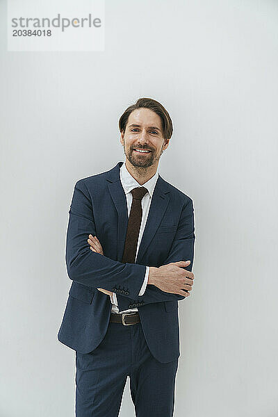 Smiling businessman standing with arms crossed in front of white wall