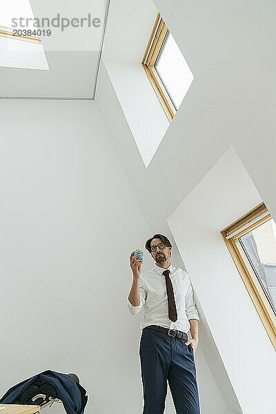 Businessman holding globe leaning on white wall at office