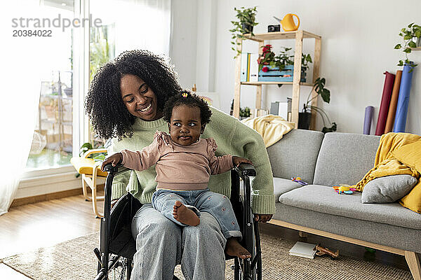 Cute baby girl sitting on young mother with disability in wheelchair at home