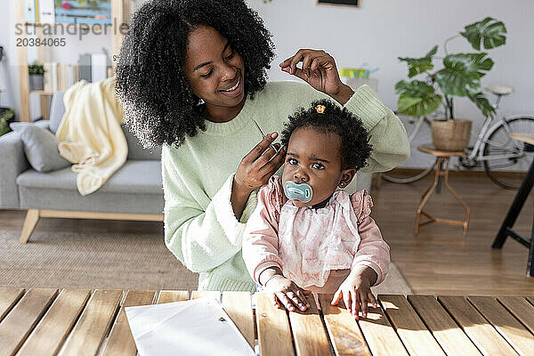 Smiling single mother with scissors cutting hair of baby girl at home