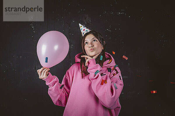 Thoughtful girl in hat holding balloon at birthday party in front of black background