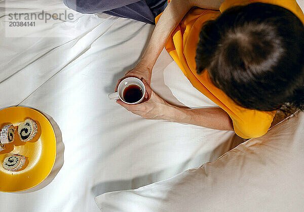 Woman in yellow t-shirt with tea cup and snack lying on bed at home