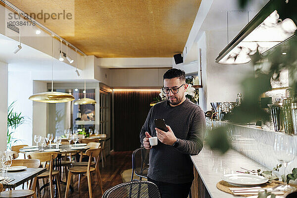 Customer using mobile phone leaning on bar counter with coffee cup at restaurant