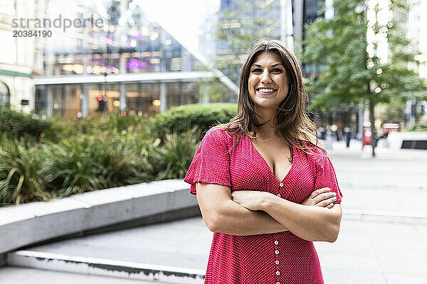 Happy young woman standing with arms crossed in front of buildings