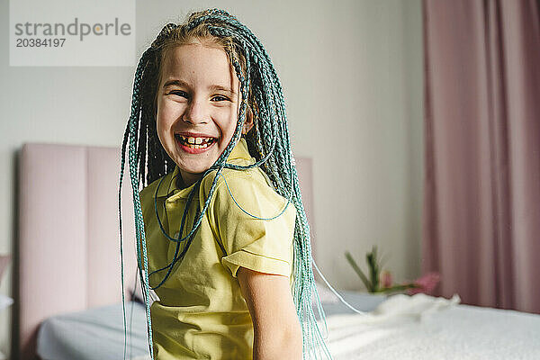 Happy girl with turquoise dyed braided hair sitting on bed at home
