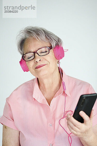 Senior woman enjoying listening to music through wired headphones against white background