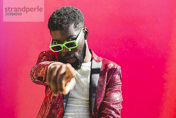 Young non-binary person wearing sunglasses and pointing against pink background