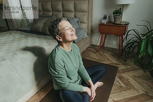 Smiling woman sitting with eyes closed near bed at home