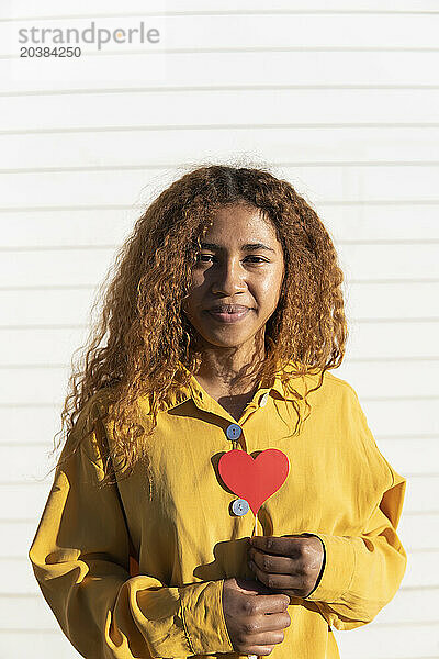 Smiling young curly haired woman standing with red heart shape prop in front of white shutter