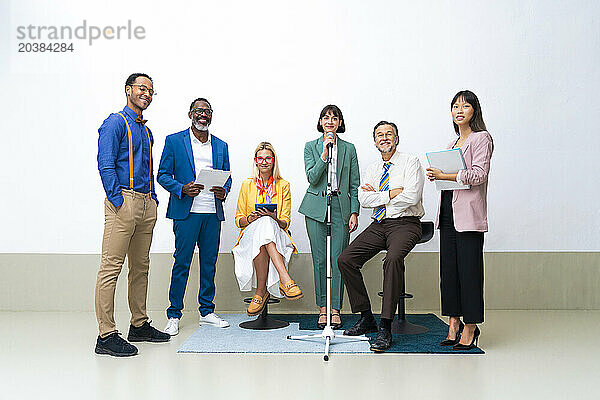 Smiling colleagues with businesswoman holding microphone near wall at office