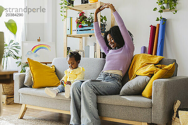 Smiling young mother stretching with hands raised sitting on sofa by daughter in living room