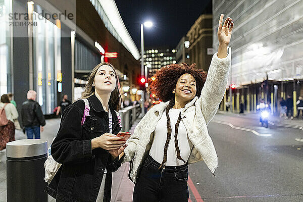 Happy woman hailing ride with friend holding smart phone on street at night