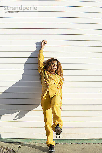 Young woman in yellow casuals leaning with hand raised on white shutter