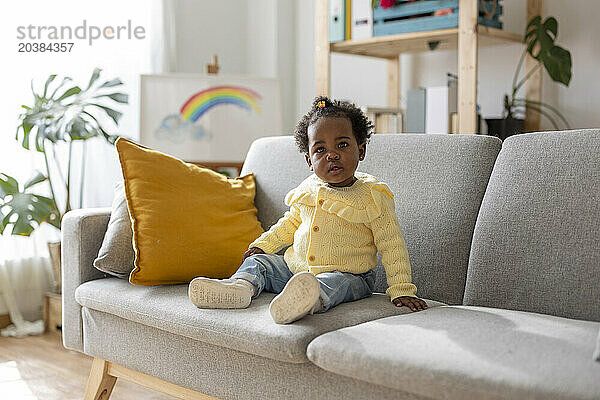 Cute baby girl sitting on sofa in living room at home