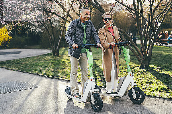 Happy couple riding electric push scooter on footpath near trees