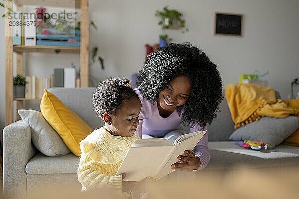 Cute baby girl and happy mother reading book in living room at home