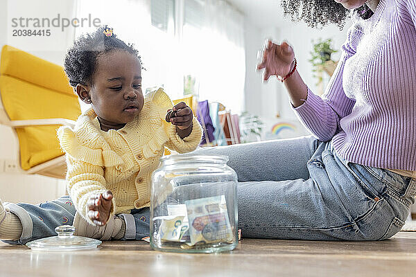 Young mother sitting near daughter playing with coins by jar of currency at home