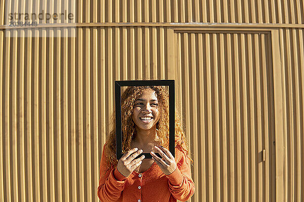 Smiling young woman looking away through picture frame standing in front of metal wall