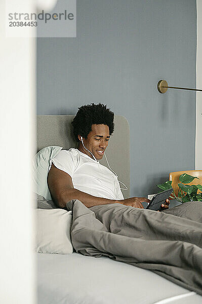 Smiling man with in-ear headphones using tablet PC on bed in bedroom at home