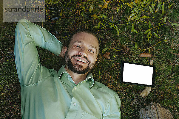 Happy businessman lying on moss near tablet PC
