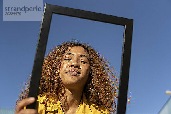 Confident woman looking through picture frame under clear blue sky