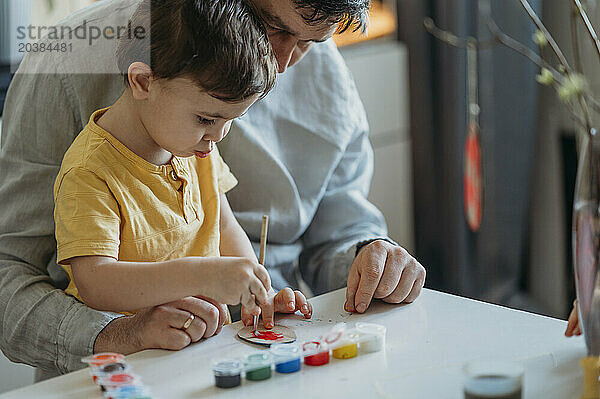 Father teaching son to paint cardboard Easter eggs with watercolors