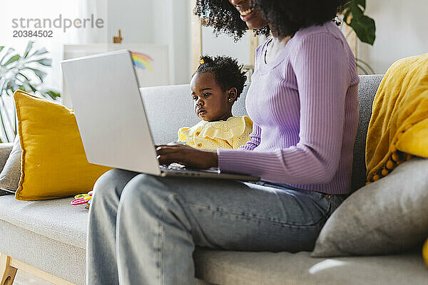 Young businesswoman using laptop with baby girl sitting on sofa in living room at home