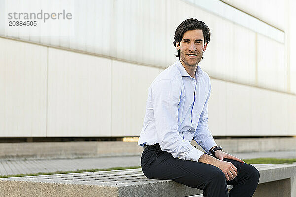 Smiling young businessman siting on wall in front of building